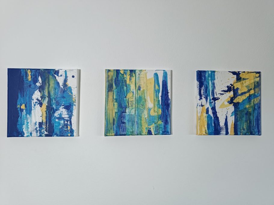 Drei quadratische Kunstwerke in Blau- und Gelbtönen an einer Wand.